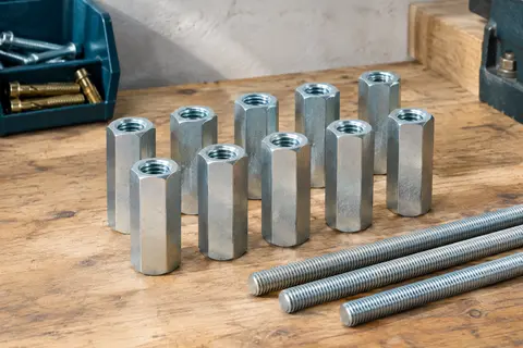Lot de dix écrous longs hexagonaux M6 zingués posé sur un établi en bois avec des tiges filetées